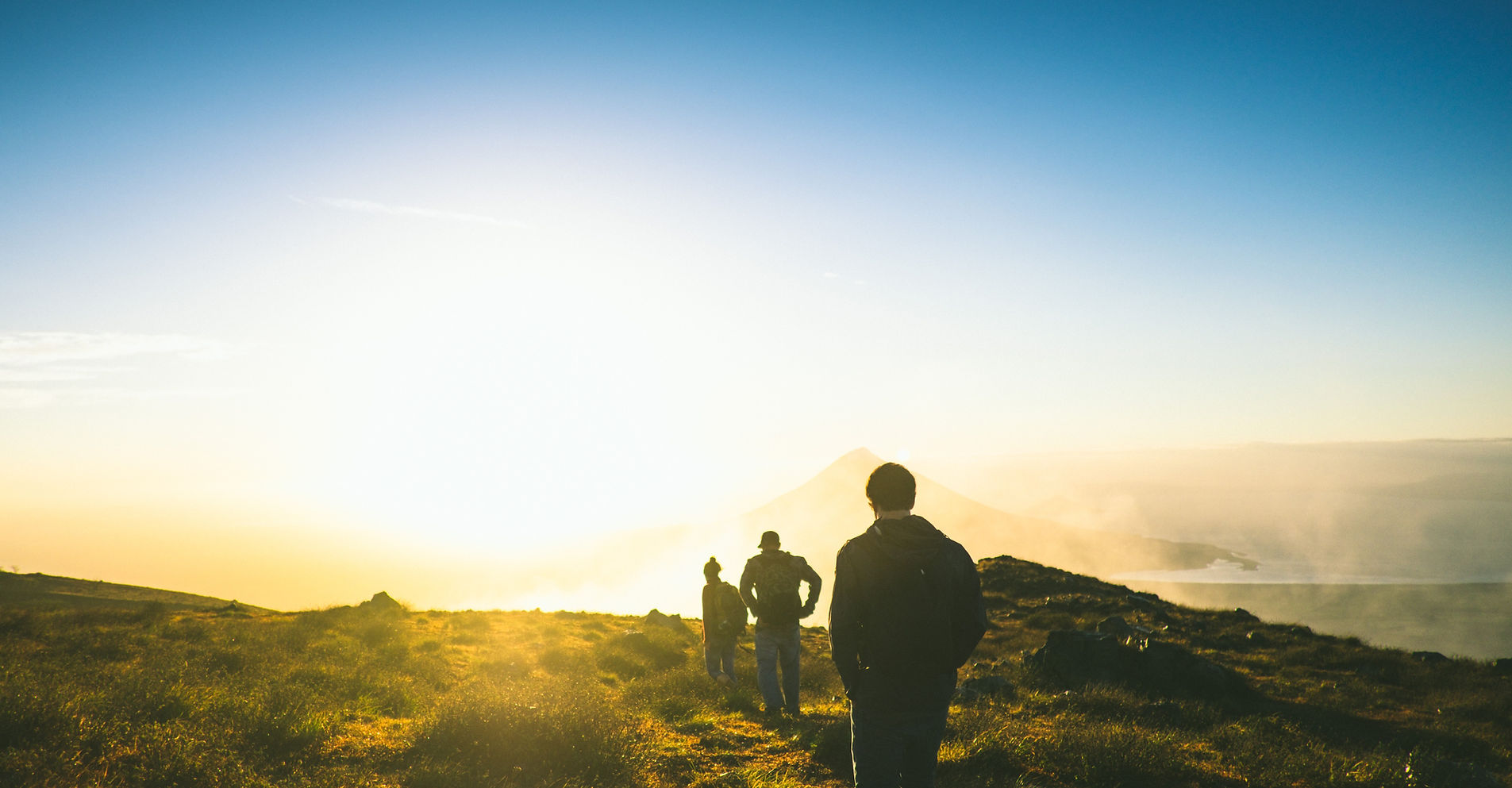 Three people walking across a grassy hilltop toward a sunlit horizon